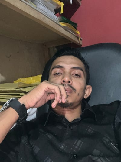 The image depicts an adult male seated indoors, located in Alueduamuka S, Indonesia. The subject, an individual with a medium skin tone, dark hair, a mustache, and a goatee, is presented from the chest up. He wears a black collared shirt featuring a subtle dark plaid pattern. A wristwatch, comprising a silver-toned metallic bracelet segment and a black strap, is visible on his left wrist. His left hand is raised, with fingers resting against his chin and lower lip. He maintains direct eye contact with the camera. The background reveals a wooden shelf unit in the upper left, containing stacks of papers and binders in yellow, white, and green colors. A partial view of a red wall is visible in the upper right background, positioned behind a dark office chair where the subject is seated. The scene is illuminated by artificial light sources.