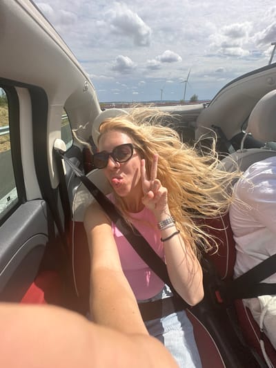 The image shows a woman in a convertible car, with her hair blowing in the wind. She is sticking her tongue out and holding up two fingers in a peace sign. She is wearing sunglasses, a pink top, and denim shorts. The car is on a road with windmills in the background. The location is Nouvion-le-Comte, France.