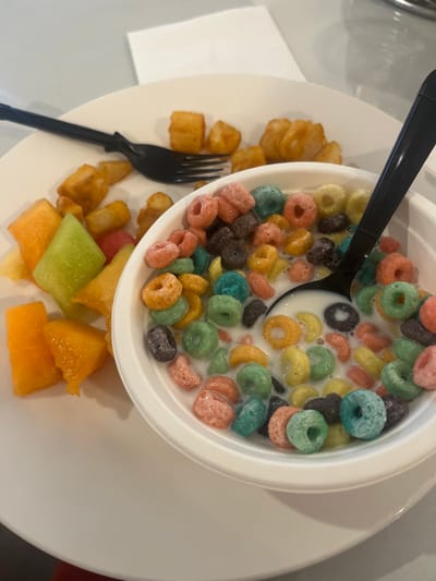 A white plate holds a bowl of colorful cereal with milk and a black spoon inside. Next to the bowl, there are golden-brown potato tater tots, a black fork, and chunks of cantaloupe and melon. The presentation suggests a combination of breakfast staples. A white napkin is visible on the left side of the plate.