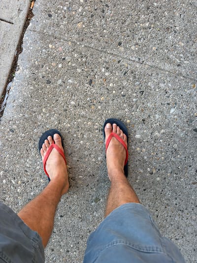 An overhead, eye-level shot shows a pair of bare feet in blue and red flip-flops, standing on a concrete sidewalk. The sidewalk has small pebbles embedded in it, and to the left, there is a grassy verge with a cigarette butt on it. The person is wearing grey shorts. The image was taken in Riverhead, United States of America.
