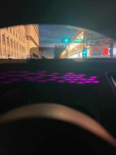 The image depicts an exterior urban night scene viewed through a vehicle's windshield, looking northbound on Virginia Street in Reno, Nevada, United States of America.

In the foreground, the vehicle's dark dashboard is visible, featuring multiple rows of small, blurred, illuminated magenta and purple lights. The upper arc of a dark steering wheel is present at the bottom of the frame.

The exterior scene reveals a multi-story classical-style building with numerous windows and columns on the left, illuminated by warm orange light. To its right and slightly behind, a taller, modern building is visible. On the right side of the street, a commercial building with several brightly illuminated signs is prominent. Visible signs include "Cal Neva - Reno's Best Bet" and "WELCOME RENO'S BEST BET" in red and white. Two green traffic lights are active: one suspended over the intersection and one mounted on a pole near the commercial building. A pedestrian signal displaying a white 'walk' symbol is visible on the left. The sky is dark blue with faint cloud formations, indicating late evening or night. The overall environment is brightly lit by commercial signage and street lighting.
