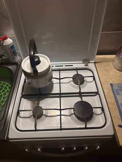 A white gas stove is shown in a kitchen, with a white enamel kettle with a red floral design placed on the left burner grate. To the left of the stove is a stainless steel sink with a green plastic drainer. A bottle of SARMA dishwashing detergent is visible next to the sink. The stove has four black burners and a white cooktop. The stove's lid is open, revealing the burners. The background shows a tiled wall and a countertop.