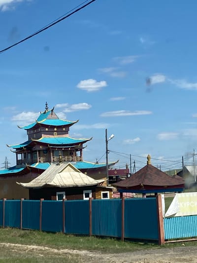 Here is a description of the image:
The image shows a vibrant and detailed architectural structure, seemingly a multi-tiered pagoda or tower, characterized by its blue and brown color scheme and ornate design elements. The architecture appears to be influenced by Asian styles, possibly Chinese or Japanese. This structure is situated next to what seems to be a modern, utilitarian building, which is constructed from blue corrugated metal sheets. The ground surrounding the buildings features green vegetation.
In the background, a clear blue sky dotted with scattered white clouds is visible. There are power lines running across the sky, suggesting the structures are situated in a developed area.
Based on the information available, this image likely depicts a site in Ust-Ordynsky, Russia. The ornate pagoda structure stands in contrast to the more functional, modern structure beside it.