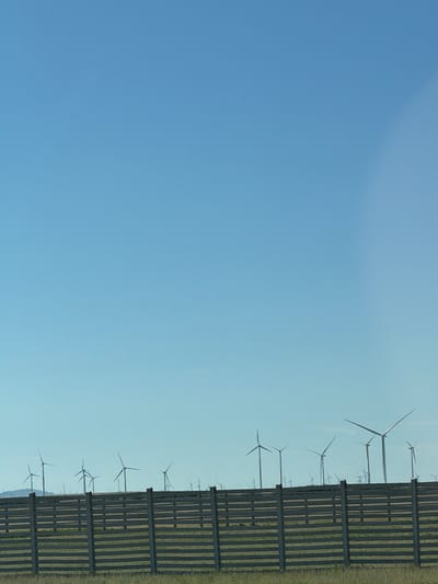The image depicts a clear blue sky over a landscape dotted with numerous wind turbines, indicating a wind farm. In the foreground, a wooden fence runs horizontally across the frame, with a grassy area visible beyond it. The wind turbines are silhouetted against the sky, with some appearing closer and more defined than others in the distance. The overall scene suggests an open, rural environment, likely in an area with consistent wind resources. The location is specified as Ereymentaw, Kazakhstan.