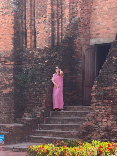 A woman in a long pink dress and sunglasses stands on the steps of an ancient brick structure in Tuy Hoa Ward, Vietnam. The structure appears to be a temple or tower, with weathered red bricks and intricate architectural details. The woman is facing to her left, with her right hand resting on her head. The steps leading up to the entrance are also made of brick, and are partially covered in moss and greenery. Red flowers bloom at the base of the structure, adding a splash of color to the scene. The sky is clear and sunny, casting shadows on the bricks.