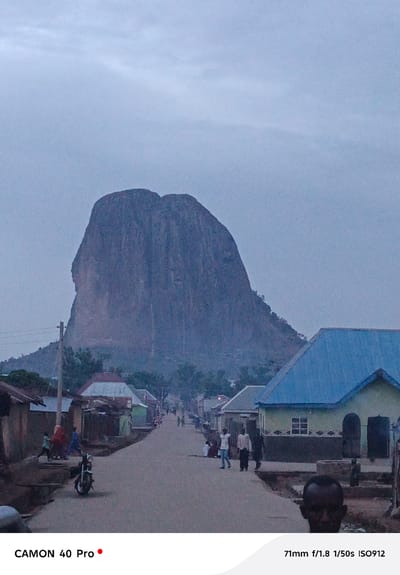The image captures a street scene in Wase, Nigeria, dominated by a large rock formation in the background, likely Wase Rock. The street is unpaved and lined with simple buildings, some with corrugated metal roofs, including one painted light green with a blue roof. Several people are visible along the street, some walking, others standing or sitting. A motorcycle is parked on the left side of the road. The sky is overcast, suggesting dusk or dawn. The presence of people and buildings, along with the prominent rock formation, indicates a rural settlement in the vicinity of Wase Rock.