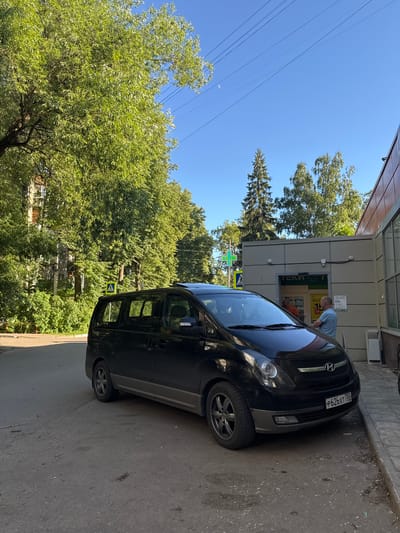 A black minivan is parked on a paved road in front of a building with a glass entrance, possibly a store or pharmacy, in Bykovo, Russia. The minivan has a license plate that reads "P626XT 750". A man in a light blue shirt is standing by the entrance, facing away from the camera. He appears to be interacting with someone inside the building. Trees and a building are visible in the background, and overhead power lines are present. A pedestrian crossing sign is also visible near the road. The sky is clear and blue, with a sliver of a moon visible.