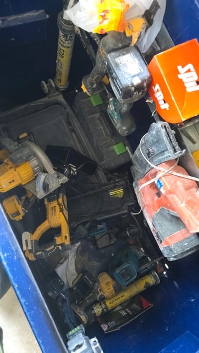 In a blue plastic container, a collection of power tools and construction supplies are stored. Among the items are a circular saw, a nail gun, several battery-powered drills, and battery packs. Some of the tools are yellow and black, consistent with the DeWalt brand, while others are teal and black, possibly Makita. There are also black plastic tool cases, a yellow sealant tube labeled "High Ratio Pu," and an orange box with the word "Spot" printed on it. The contents appear to be dusty and possibly used. The context of Milton Keynes, United Kingdom, is not discernible from the image itself, as it lacks any identifying landmarks or signage.