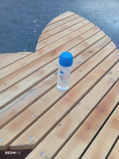 A clear plastic bottle of water with a blue cap and a label featuring a stylized blue drop and the word "RARE" is positioned on a light-brown wooden slatted surface. The surface appears to be part of outdoor seating or a bench, with visible screws securing the slats. The background consists of dark asphalt or pavement with scattered small stones and debris. The image is cropped to focus on the bottle and the wooden surface, with the lower left corner displaying the text "REDMI 12." The scene does not show any people or specific city landmarks, but the information provided about the location in Vedi, Armenia, suggests an outdoor setting within that city.
