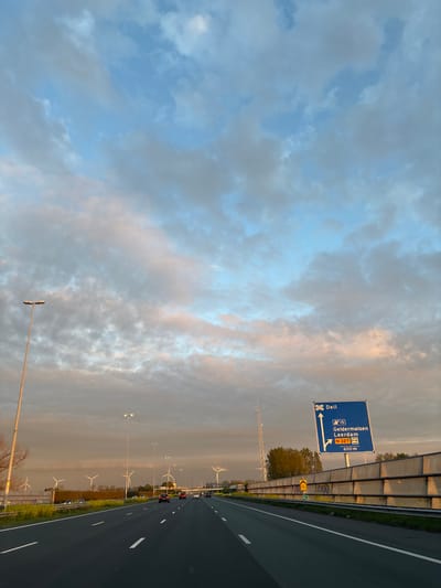 The image depicts a highway in the Netherlands, likely near Beesd given the road sign pointing towards Deil, Geldermalsen, and Leerdam. The sign indicates directions for the A15 and N327 routes. The road is divided with multiple lanes in each direction, and some traffic is visible. Wind turbines are sparsely distributed on the flat landscape adjacent to the highway. A noise barrier runs alongside the road. The sky is partly cloudy, with the setting sun casting an orange hue on some of the clouds.