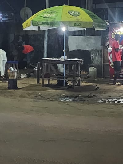 The image depicts a street scene in Obiaruku, Nigeria. The focus is on a simple outdoor eating area with wooden tables and chairs, partially covered by a large, advertising umbrella for the GLO telecommunications company. People are present in the background, though details are obscured by shadow and distance. The ground appears to be a mix of dirt and pavement. The atmosphere seems to be low-light, possibly evening. There is no indication of any interactions occurring in the image.