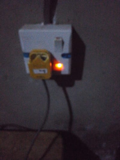 A white power adapter is plugged into an outlet. A yellow plug is inserted into the adapter. An orange light indicates power. The adapter has a switch and a blue stripe. Black cords extend from the yellow plug downward. The background is a dark, grainy wall.