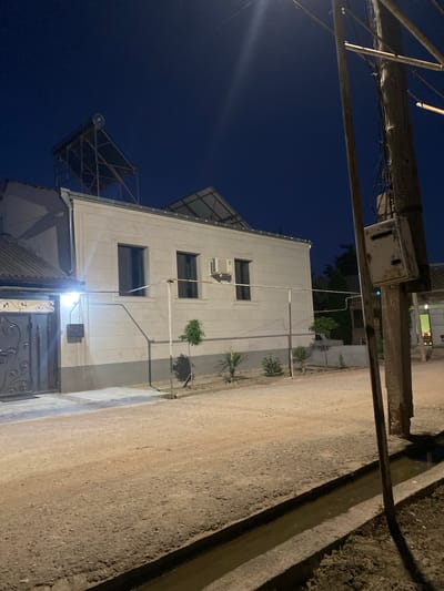 The image shows a three-story building at night, likely in Verin Artashat, Armenia. The building is made of light-colored material and has several windows. An air conditioning unit is visible on the second floor. There are solar panels on the roof, indicating a focus on renewable energy. A metal gate is in front of the building. The ground is a mixture of dirt and gravel, with a concrete structure running alongside it. A utility pole with wires is also visible. The sky is dark blue, suggesting it is nighttime. The lighting suggests artificial illumination from the building and possibly streetlights. There are some small, spiky plants in front of the building. The overall impression is one of a residential or commercial building in a somewhat rural or developing area. There is no visible text in the image.