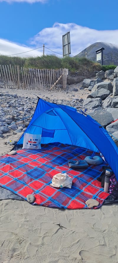A blue beach tent is set up on a sandy beach in Pier Road, Ireland. The tent is open, revealing a red and blue plaid blanket laid out inside. On the blanket, there are two inflatable pillows, a gray bag, a white fabric bag with a logo, and a metal thermos. Several stones are scattered around the blanket and the entrance to the tent. In the background, a wooden fence lines a grassy area, and large rocks are piled along the coastline. Beyond the rocks, a hill with a misty peak is visible under a partly cloudy sky. The sand in the foreground is marked with numerous footprints.