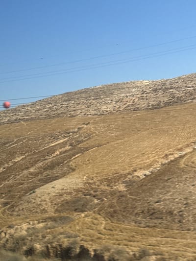 The image depicts a rocky, arid landscape with a clear blue sky. The scene is dominated by a large, textured rock face that fills the right side of the frame. The rock appears weathered and has a stratified appearance, with shades of brown and beige. Several thin, dark lines, likely power lines or wires, run diagonally across the left side of the image, against the sky.
In the lower left portion of the image, a small, bright pink sphere is visible, hanging from one of the wires. Further up and to the left, a tiny, lighter-colored object, possibly a small balloon or drone, is also suspended in the sky. The overall impression is one of an outdoor scene, possibly in a remote or natural setting. The lighting suggests it is daytime, and the clear sky indicates fair weather. There is no visible text in the image. The context provided suggests this scene is located within the al-Qasoum Regional Council in Israel.
