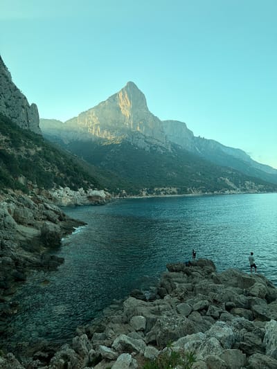The image shows a dramatic coastal landscape with steep, rocky cliffs overlooking a vast expanse of blue-green water. The sun is setting, casting a warm golden light on the mountains in the background, creating long shadows and highlighting the rugged textures of the terrain. The sky is a clear, pale blue, indicating fair weather.

In the foreground, large, weathered rocks form the edge of the cliff. Several people are visible on these rocks, near the water's edge. One person is standing, looking out at the sea, while another appears to be in the process of jumping or diving into the water. Another person is seated on a rock further down. The water is calm, with gentle ripples reflecting the sky and surrounding cliffs. The overall mood is one of awe and adventure, with a sense of peaceful solitude mixed with the thrill of extreme sports.

The scene is set in a remote, natural environment, likely a popular spot for cliff diving or swimming. The vegetation on the slopes suggests a Mediterranean climate. The orientation of the image is vertical, giving a sense of height and depth. There is no visible text in the image. The location context of Baunei, Italy, is consistent with the depicted scenery of rugged coastline and clear waters.