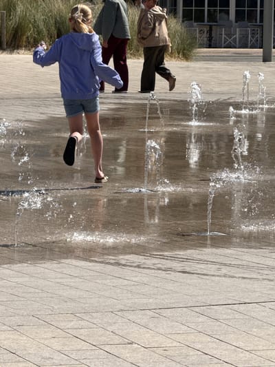 The image captures an outdoor public space in Middelkerke, Belgium, featuring an interactive water fountain under bright sunlight. The ground is paved with light-colored stone or concrete tiles arranged in a diagonal pattern.
Three individuals are visible. In the foreground, a person with blonde hair, wearing a light purple long-sleeved top and light blue shorts, is actively engaging with the water jets. They wear black and light-colored sandals, with one foot lifted as if running or stepping through the splashing water. Their back is towards the camera. To the left, another person in a light brown jacket and dark pants walks away, partially obscured. A third individual, wearing a dark green top and maroon pants, is partially visible behind the person in purple.
Numerous streams of water erupt vertically from the ground, creating splashes and reflections on the wet surface. The strong sunlight illuminates the water, making it sparkle, and casts clear shadows from the people, indicating a sunny day. In the background, a modern building with large windows is visible. Faint text, possibly "CAFE", can be seen through one of the windows, although it is somewhat indistinct. The scene conveys a lively, pleasant atmosphere.