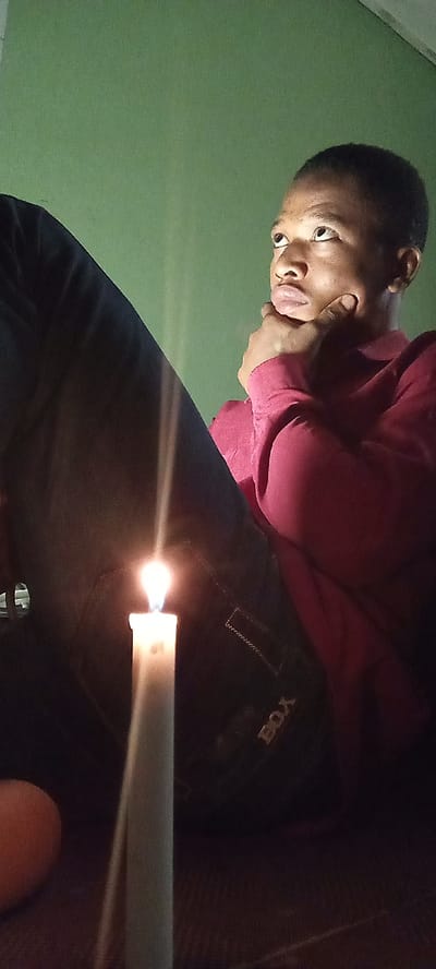 A young man is seated with his legs crossed, illuminated by a candle in a dimly lit room. He is wearing a red long-sleeved shirt and dark pants with the word "BOX" visible on the back pocket of his pants. His right hand is raised, with his chin resting on his fingers. He is looking up and to the left, seemingly in thought. The candle is in the foreground, providing the primary light source. The wall behind him is a pale green color. The location is presumed to be Oshogbo, Nigeria, based on external information not evident in the image.