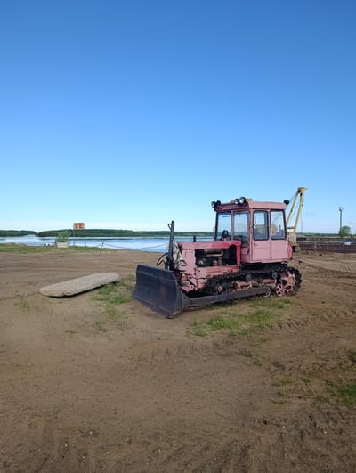 The image displays an outdoor scene set on a sandy riverbank or industrial area under a vast, clear blue sky. The ground in the foreground is predominantly sand or light dirt, interspersed with small patches of green grass.

The central subject is an aged, tracked bulldozer, painted a faded pink or reddish hue, positioned slightly right of center. It features a robust black front blade, an exposed engine compartment, and a driver's cabin with glass windows. Four distinct round lights are mounted on the cabin's roof. The bulldozer's tracks show signs of wear and rust. To the left of the bulldozer, a long, rectangular concrete slab lies flat on the ground.

In the middle distance, a wide, tranquil body of water, likely a river or lake, extends across the frame, bordered by a dense, green forested bank on the opposite side. Near the water's edge on the closer bank, a rusty, rectangular metal sign or structure is visible, partially hidden by a small green bush.

Further back and to the right, a yellow crane arm is partially discernible behind the bulldozer. A dark, low-lying barge or dock structure is moored on the water, with faint white Cyrillic text visible on its side, which appears to read "ОГРАНИЧЕНИЕ ПРИЧАЛ" (Restriction Pier/Dock). A tall, black pole with a light fixture stands near this structure.

The weather is bright and sunny, characterized by the cloudless blue sky. No people are present, and the scene depicts a static, idle moment, possibly at an industrial or port area in Igrim, Russia.