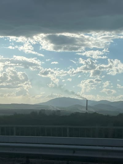 The photograph shows a view from a vehicle, likely a car, of the landscape near Balanovo, Bulgaria. In the distance, there is a mountain range with a tall industrial chimney or tower visible, emitting what appears to be smoke or steam. The sky is filled with clouds, ranging from dark grey to bright white, suggesting a mix of weather conditions. The foreground features a series of metal posts or a barrier, indicating the photo was taken through a window.