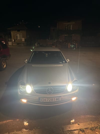 At night, a silver Mercedes-Benz CLK is parked on a paved surface that appears wet, likely from rain. The car's headlights are on, illuminating the front of the vehicle and the ground in front of it. The license plate reads "EH 7281 ME" with "BG" indicating Bulgaria. In the background, there are buildings, one with a red roof, and stacks of firewood. A red off-road vehicle is partially visible to the left of the Mercedes. The scene is likely in the city of Shipkovo, Bulgaria.