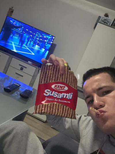 A man in a grey shirt is holding a package of "Susamli" sesame sticks, eating one, while sitting in a room with a television displaying a blue image. The room includes a table with two black objects on it, and a white cabinet. There is no information to suggest that the scene is in Benningen, Germany.