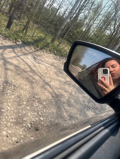 A woman is reflected in a car's side mirror while taking a photo of herself with a white cell phone. The exterior of the car is gray. The mirror reflects a rural scene of a dirt road lined with trees. The road's surface is light gray with scattered stones. The trees are thin and have light-colored trunks. The image was taken in Cheryomushki, Russia.