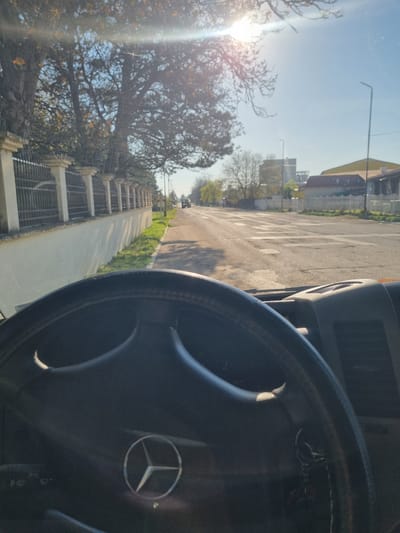 The image is taken from inside a vehicle, showing a Mercedes-Benz steering wheel. Outside the windshield, a street scene unfolds in Ovcharovo, Bulgaria. Buildings are visible along the street, with trees and foliage lining the sidewalk. The street appears to be in a residential area, and the sun is shining brightly.