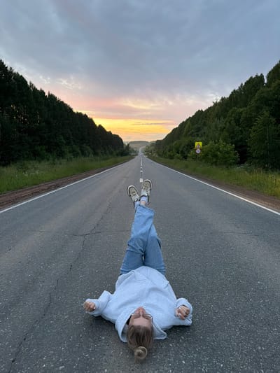A person lies supine on an asphalt road, which is flanked by dense green trees on either side. The road extends into the distance, where the horizon is marked by a pale orange and pink sunset. The person is wearing a light gray hooded sweatshirt, blue jeans, and sneakers. Their legs are bent and crossed at the ankles, and their arms are outstretched to their sides. A speed limit sign with a "70" is visible on the right side of the road. The sky is overcast with soft clouds, and the overall scene suggests a rural setting.