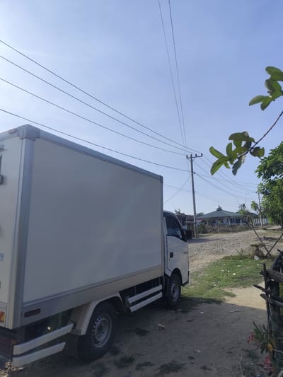 The image captures a static outdoor scene in Cotgirek, Indonesia, on what appears to be a bright, sunny day with a clear blue sky. The primary subject is a white box truck, seen from its right side, parked on a dirt and gravel path. The truck features a large, plain white cargo box with silver-colored trim on its edges. Part of the cabin is visible, and the word "TRASSA" can be faintly read on its side. Near the rear wheel well, a yellow triangular sticker and partially obscured text, possibly "VAC...", are visible.
Several power lines stretch horizontally across the sky, originating from a utility pole located further down the path on the right side of the frame. Beyond the utility pole and power lines, a building with a distinctive green-domed roof, characteristic of a mosque, is visible in the middle distance. Green foliage, likely from a tree or shrub, partially frames the right side of the image, with some leaves in the foreground appearing slightly out of focus. The ground is a mix of packed dirt, loose gravel, and sparse patches of green grass. No specific activity or people are depicted; the scene suggests a quiet, possibly rural or semi-rural setting. The bright sunlight casts some shadows, indicating a time of day with strong overhead light, possibly late morning or early afternoon.