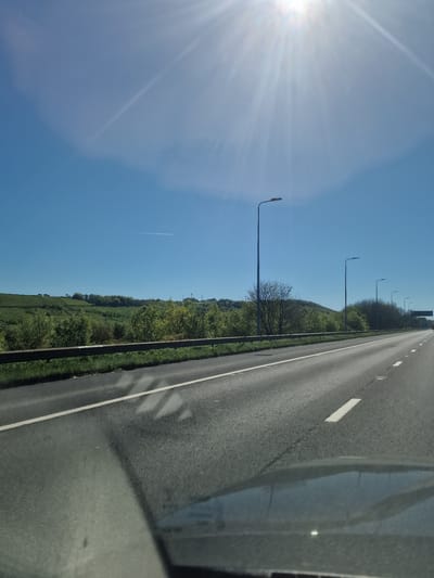 The image depicts a bright, sunny day on a motorway or major highway, likely taken from inside a moving vehicle in Kirklees, United Kingdom. The perspective looks forward and slightly to the left, with the multi-lane road occupying the right side of the frame, receding into the distance. White road markings and reflective studs are visible on the asphalt.
To the left of the road, a narrow grassy verge leads to a series of tall, slender streetlights. Beyond these, a dense line of green trees and bushes forms a barrier, behind which a expansive, undulating landscape of vibrant green fields and rolling hills stretches towards the horizon. The sky is a clear, light blue, dominating the upper portion of the frame. A faint, white contrail from an aircraft is visible high in the sky above the landscape.
The sun is a prominent feature in the upper-left corner, emitting a strong lens flare and bright rays that spread across the sky, highlighting the clear weather conditions. On the far right, the dark interior of the vehicle, possibly a window frame, is visible, along with reflections on the glass showing distorted shapes of the road and surrounding elements. The scene conveys a sense of open-road travel through a picturesque, rural or semi-rural environment under brilliant daylight. No people or legible text are visible.