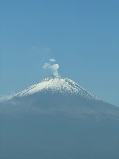 An image of a snow-covered volcano emitting a plume of smoke against a clear blue sky. The scene is in the city of San Bernardino Chalchihuapan, Mexico.