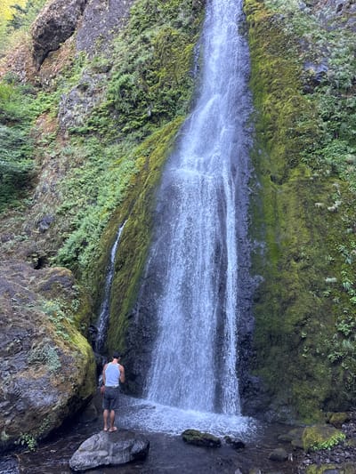 The image displays Wyeth Falls in Wyeth, Oregon, United States. A tall, multi-tiered waterfall descends over a steep rock formation heavily covered in green moss and other vegetation, emptying into a shallow pool at its base. One adult male is present in the foreground, standing barefoot on a large, dark-colored boulder situated within the water. He is viewed from behind, wearing a white sleeveless shirt and dark shorts, and is facing the waterfall. The immediate environment includes additional large, moss-covered boulders and dense green foliage, consistent with a lush, wet natural setting.