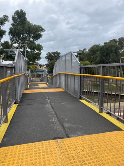 The image depicts a pedestrian overpass at Bassendean railway station in Bassendean, Western Australia. The overpass has metal mesh sides and a bright yellow handrail. A black asphalt pathway runs down the center of the overpass, bordered by a yellow safety strip on the right side. The right side also features a tactile ground surface indicator with a yellow bumpy texture. Beyond the metal mesh sides are trees and a glimpse of buildings. The sky is overcast.