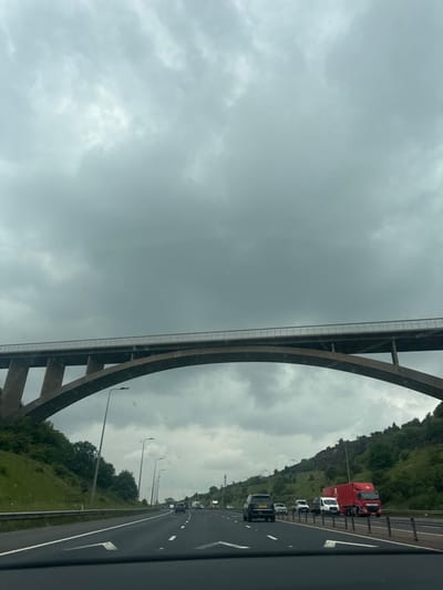 The image shows a view from inside a car driving on a highway. The car is driving under a large concrete bridge with a slight curve. The bridge is high above the highway. There are a few cars on the highway, including a red truck with a white trailer on the right side. The highway is lined with green vegetation. The sky is overcast, and the image is taken on a day with a hint of sun. The view is from the M62 motorway at the location of the Junction 26, which is located in the city of Kirklees, United Kingdom.