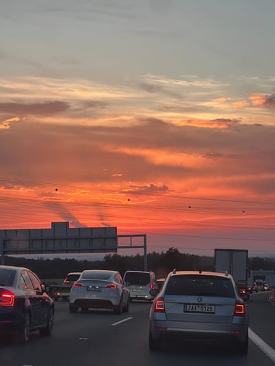This is a photograph taken from a car, likely in traffic, on a highway in Všechromy, Czechia. The image is oriented vertically, with the sky filling the left side and the road and vehicles on the right. The sky is a dramatic sunset, with vibrant orange, pink, and yellow hues reflecting off clouds. Power lines are visible stretching across the sky.
On the road, several cars are stopped or moving slowly. The most prominent vehicle is a silver hatchback with its taillights illuminated. Its license plate is clearly visible and reads "7AX 0125". Further down the road, other vehicles, including trucks and cars, are visible in the distance. The presence of traffic suggests an event like rush hour or a traffic jam.
The overall mood is peaceful, perhaps even awe-inspiring due to the beautiful sunset. The time of day is clearly sunset. The location is identified as Všechromy, Czechia, which aligns with the appearance of the license plate. There are no people visible in the photograph. The scene is set on a highway, indicated by the road markings and the presence of multiple vehicles.