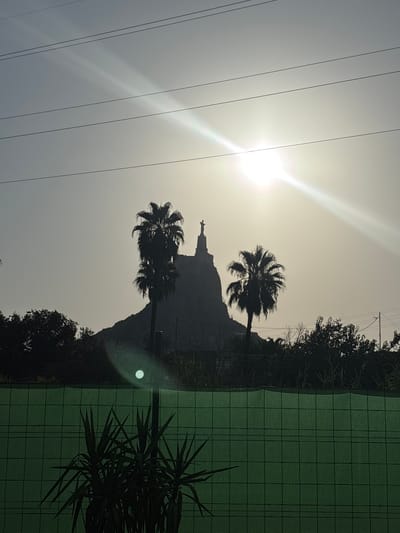 The image captures a silhouetted view of a statue atop a rocky hill, flanked by two palm trees, against a bright, hazy sky. The sun is partially obscured by the haze, creating a strong lens flare and a bright glow. Multiple power lines crisscross the sky above the scene. In the foreground, a dark green fence with a grid pattern is visible, with some dark green foliage, possibly a yucca plant, in front of it. The overall scene suggests an outdoor setting, likely in a warm climate, possibly during the late afternoon or early morning due to the low angle of the sun and the hazy light. The statue atop the hill appears to be a religious figure, possibly a cross or a statue of Jesus Christ, given its common placement on prominent hills in Spain. This specific location is identified as Cabezo De Torres, Spain, a municipality in the autonomous community of the Region of Murcia. The silhouette of the statue, the palm trees, and the hilltop are characteristic of religious monuments often found in Spanish towns.