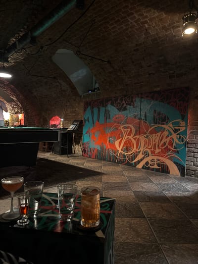 The image depicts the interior of a bar with a low, arched brick ceiling. A large, vibrant graffiti artwork is prominently displayed on a brick wall. In the foreground, a tray holds several drinks, including a cocktail glass, two shot glasses, and two highball glasses. To the left, a portion of a pool table is visible. The floor is tiled with dark, square tiles. The overall ambiance suggests a casual, artistic, and possibly dimly lit establishment. There are no individuals present in the scene. The location of Novaya Usman, Russia, is not discernible from the visual content of the image.