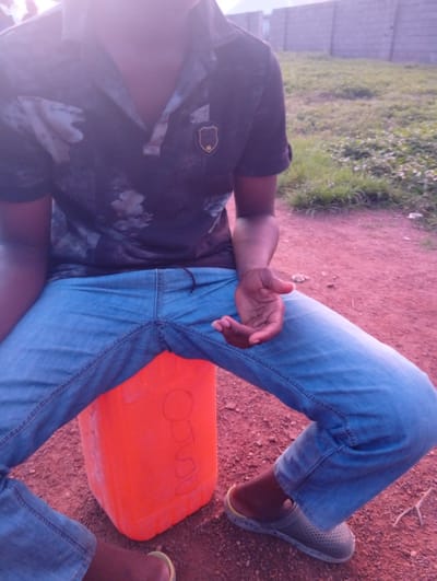 A person is sitting on an orange plastic container in an outdoor environment, possibly in Balewa, Nigeria. The individual is wearing a dark patterned short-sleeved polo shirt and blue denim jeans. Their right hand rests on their lap. The ground is a reddish-brown dirt or gravel, with patches of dry grass and sparse vegetation in the background. A grey brick wall is visible in the distance, suggesting a built-up area or a boundary. The lighting suggests it is either early morning or late afternoon. The overall scene is informal and casual.