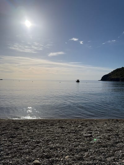 The image depicts a tranquil coastal scene at a pebble beach in Colera, Spain. The sun is high in the sky, casting a bright glare and creating shimmering reflections on the calm, blue water. The sea stretches towards a distant horizon where it meets a pale, cloud-streaked sky. In the middle of the water, a small, dark boat is anchored. To the right, a tree-covered hillside slopes down to the sea. The foreground is dominated by a dense covering of grey pebbles that form the beach.