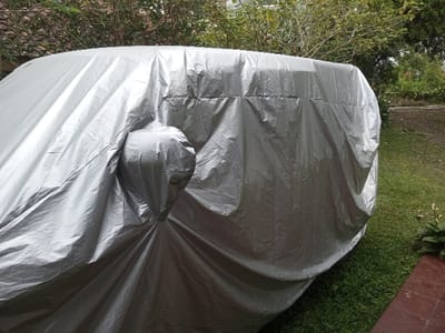 The image depicts a vehicle completely covered by a wrinkled, silver-grey protective tarp, parked outdoors in a grassy area in Naya Andura, India. A distinct protrusion on the left side of the covered vehicle suggests a side mirror beneath the tarp. The ground consists primarily of green grass, with a small section of reddish-brown pavement or tile visible in the bottom right foreground, alongside some small white flowers. The background features dense green foliage and tree branches. The scene is illuminated by natural daylight. No individuals or active interactions are present.