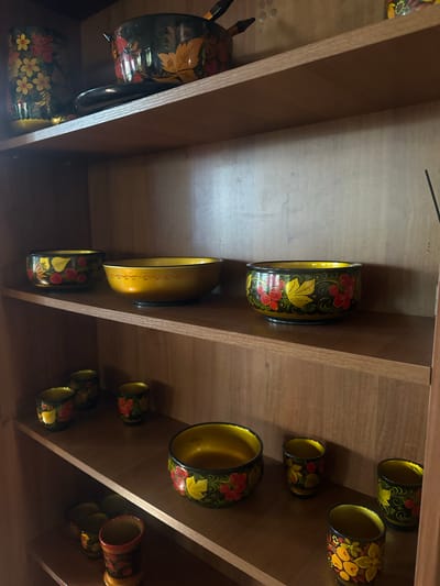 This image shows a collection of traditional Russian Khokhloma painted woodenware arranged on wooden shelves. The items include several bowls and cups, all featuring intricate floral and berry designs in red, yellow, black, and green. The objects are well-lit, with some natural light illuminating the scene from the left. The background consists of wooden shelving and the interior of a cabinet or room. There are no people visible in the image. The specific location within the city of Konstantinovka, Russia, is not identifiable from the visual information provided in the image.