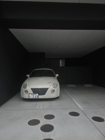 A white Daihatsu Copen convertible is parked in a covered parking garage. The car has a black honeycomb grille and a license plate that reads "Nagoya 593" and "む・3 69". The parking garage has a concrete floor with several circular manhole covers and white parking lines. The walls of the garage are dark gray.