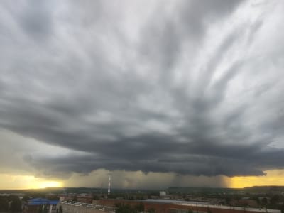 A wide-angle, high-angle view captures a dramatic sky over the city of Vyatskiye Polyany, Russia. Dark, heavy storm clouds dominate the upper portion of the frame, with shafts of rain visible beneath them, particularly in the center and to the right. The setting sun peeks through the clouds on the left, casting a warm, yellow glow and illuminating the rain streaks. Below the storm, a cityscape unfolds with various buildings, including what appear to be residential structures and industrial facilities. Two tall smokestacks are visible, one with red and white markings, amidst the urban landscape. The overall scene conveys a sense of impending weather and a contrast between the stormy sky and the illuminated horizon.