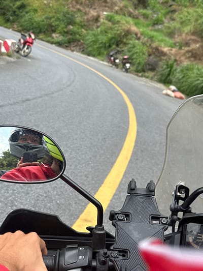 The image provides a first-person perspective from a motorcycle, looking down a winding asphalt road in what appears to be Minh Sơn Commune, Vietnam. The main subject in the immediate foreground consists of parts of a motorcycle: a clear windshield, dashboard with faint gauges, a black phone mount (currently empty), and the right handlebar with control switches. The rider's right hand, wearing a red sleeve, grips the handlebar.
Reflected in the circular rearview mirror on the left, the person taking the photo is visible, wearing a red top and holding a smartphone to capture the image. The road ahead curves sharply to the left, marked by a prominent, continuous double yellow line indicating a no-passing zone. To the left of the road are lush green grassy slopes and hills.
Further down the curved road, another red motorcycle, possibly a touring or sport bike, is visible on the left side near a red and white barrier. Near this motorcycle, a person is standing or walking alongside the road. The overall setting suggests a scenic, hilly, or mountainous area. The lighting indicates it is daytime, likely a clear day. No specific emotions are discernible, but the scene conveys a sense of a journey or a stop during a ride. Visible text on the motorcycle dashboard is too indistinct to read, apart from a possible "UP" or symbol.
