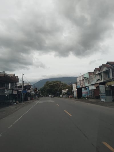 The image depicts a street in Reuhattuha, Indonesia, under an overcast sky. The road has two lanes separated by a yellow dashed line. Buildings line both sides of the street, varying in height and construction style. A mountain is visible in the distance, partially obscured by low-lying clouds. Several cars are present on the road. There are advertisements visible along the street.