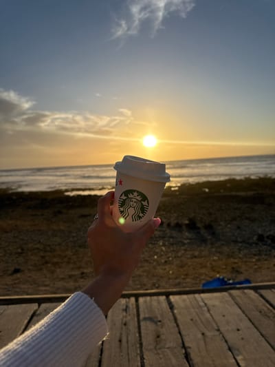 A woman holds a Starbucks coffee cup against a scenic backdrop. The sun shines brightly over the water. To the right, a wooden boardwalk runs parallel to the scene. The beach and ocean extend into the distance under a sunny sky with clouds.