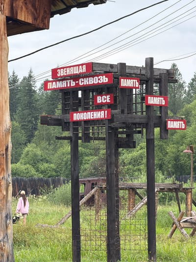 The image depicts an outdoor scene in Ust'-Kabyrza, Russia, featuring a weathered wooden structure with multiple red signs bearing white Cyrillic lettering. The signs appear to be historical or memorial in nature, with phrases like "ЗВЕНЯЩАЯ ПАМЯТЬ-СКВОЗЬ ПОКОЛЕНИЯ!" (Ringing Memory Through Generations!) and "ПАМЯТЬ" (Memory) visible. The structure is set against a backdrop of lush green trees and vegetation. In the foreground, a young person wearing a pink hoodie and shorts walks away from the camera, carrying a dark bag. The ground is covered in tall grass and wildflowers. In the background, there are remnants of what looks like an old vehicle and a rustic wooden fence. The overall atmosphere is one of historical reflection amidst a natural, rural setting.