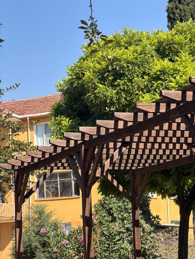 An exterior, eye-level, medium shot captures a sunlit scene with a wooden pergola in the foreground. The pergola features dark brown, weathered wooden beams forming a lattice-like roof, casting sharp shadows. Behind the pergola, a bright yellow, two-story building with a reddish-orange tile roof is visible. The building has several windows, some with white frames and curtains, and one with a rusted metal frame. Lush green trees and bushes surround the building, with a tall evergreen tree visible on the right. Pink flowers are blooming in the lower right portion of the frame. The sky is a clear, bright blue. The image appears to be taken in a residential area, possibly a backyard or garden, in Köprübaşı, Turkey. No people are present in the scene.
