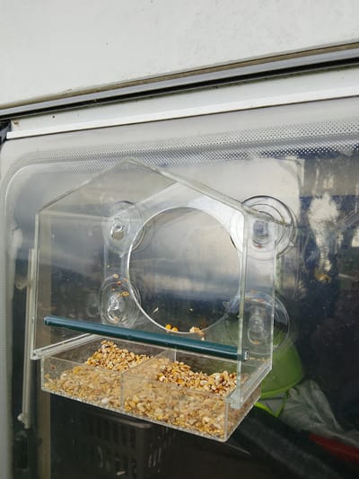 The image shows a clear plastic bird feeder, filled with birdseed, attached to a window with suction cups. The feeder has a green perch across the top. The window appears to be on a vehicle, possibly a caravan or RV, given the white exterior panel visible at the top of the image and the black material and green bucket seen through the window. The location is described as Glounaglough, Ireland. The scene is outdoors, and the lighting suggests it is daytime. There are no people or animals visible in the image.